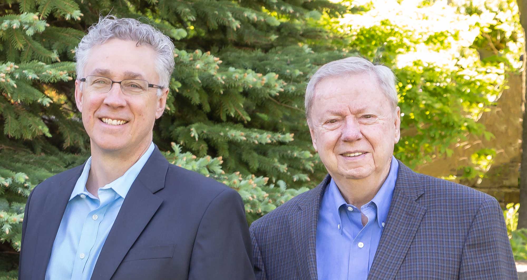 Two men standing outside smiling in the summer sun, in front of a tree.