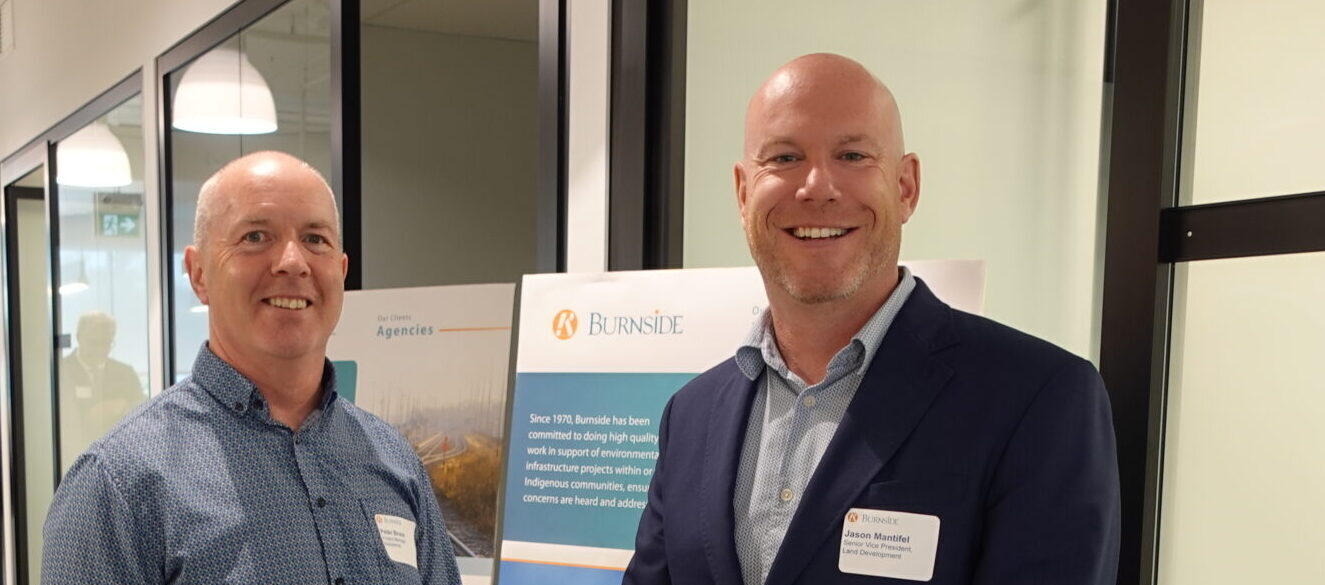 Two men standing in an office in front of a sign smiling