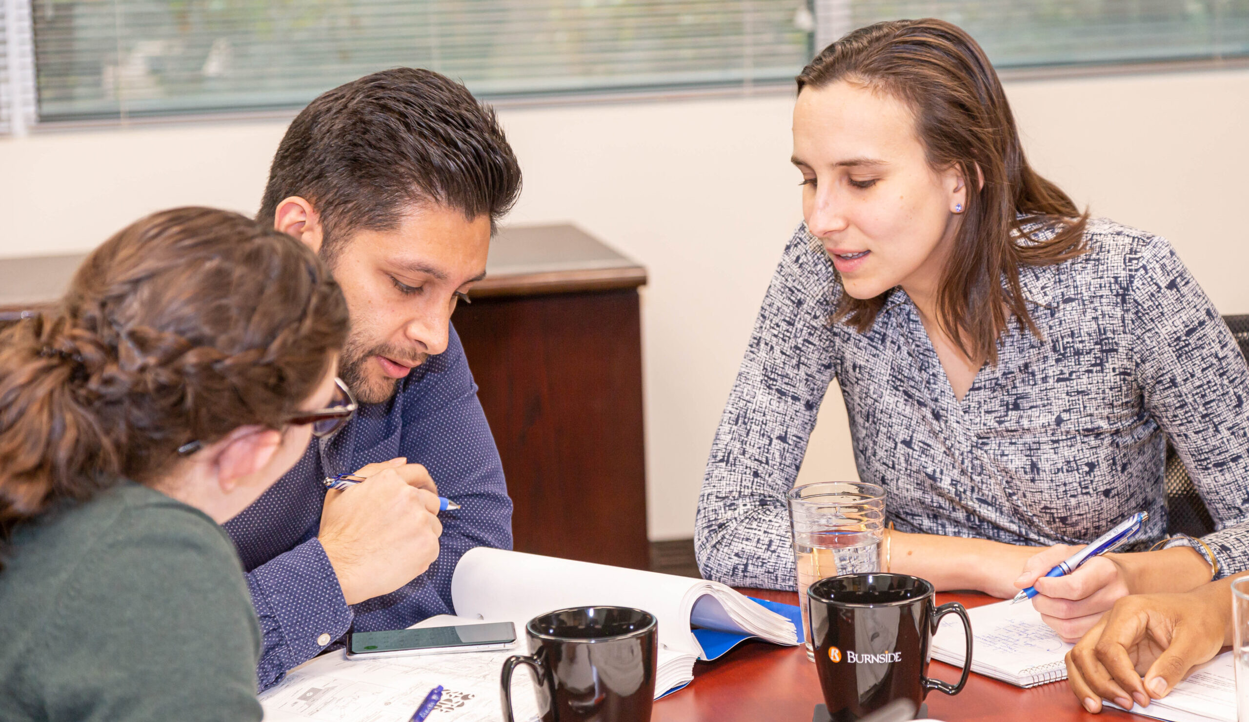 Three workers sitting at a desk looking at a book together.